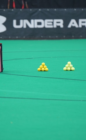 A sports field features a black and pink sports bag, two pyramids of yellow balls, and a portion of a person in dark athletic gear. The background displays a banner with the Under Armour logo.