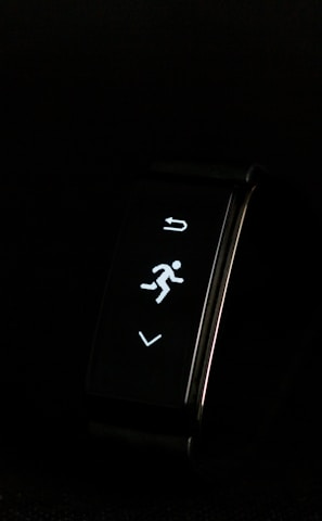 A digital fitness tracker with a black band is displayed against a dark background. The screen features minimalistic white icons for navigation and an exercise mode, including an arrow, a running figure, and a downward-pointing arrow.