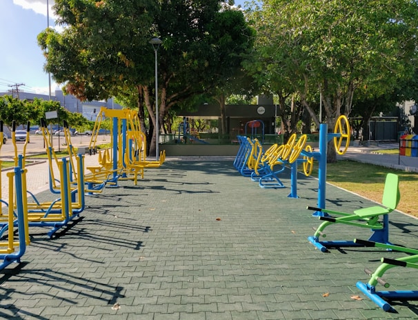 An outdoor fitness area featuring a variety of exercise equipment, including stationary bikes and other workout stations. The equipment is primarily blue and yellow, arranged neatly on a paved surface. Surrounding the area are trees providing shade, with a few parked cars and buildings visible in the background.
