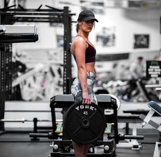 A woman is standing in a gym holding a heavy barbell plate marked with 'YORK 45'. She is wearing a cap, sports bra, and patterned leggings. The gym is equipped with various fitness machines and weights.