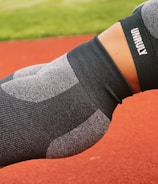 A person is performing a plank exercise on a red track field. They are wearing a matching athletic outfit consisting of a grey and black sports bra and leggings, with the word 'UNRULY' printed on the top. The background includes a blurred view of green grass.
