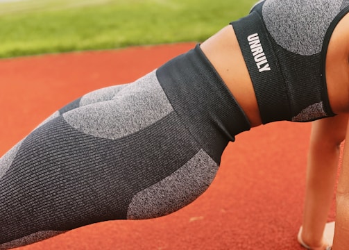 A person is performing a plank exercise on a red track field. They are wearing a matching athletic outfit consisting of a grey and black sports bra and leggings, with the word 'UNRULY' printed on the top. The background includes a blurred view of green grass.