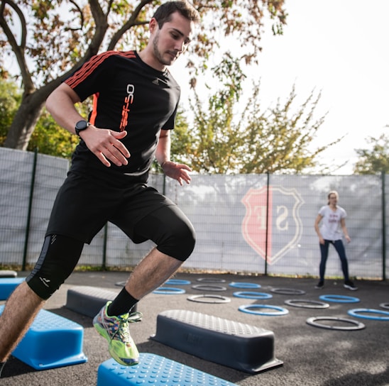 A man is actively participating in an outdoor fitness session, jumping over blue platforms with agility. Two other people in the background are also engaging in exercise, with rings scattered on the ground. The area is fenced and surrounded by trees, suggesting a park or recreational setting.