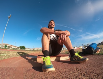 A person wearing a black athletic outfit is seated on a wooden platform in an outdoor sports field. The individual is relaxed, with arms resting on knees. The field is expansive with a clear blue sky and scattered clouds above. A brightly colored running shoe and athletic gear are visible, along with a water bottle and a blue backpack placed beside the person. Lampposts and residential buildings can be seen in the background.