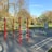 An outdoor fitness park surrounded by trees features various exercise stations with equipment like bars and rings. The area is bordered by lush green grass and there are tall trees with autumn-colored leaves in the background. The sky is clear and blue, creating a serene environment ideal for workouts.