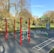 An outdoor fitness park surrounded by trees features various exercise stations with equipment like bars and rings. The area is bordered by lush green grass and there are tall trees with autumn-colored leaves in the background. The sky is clear and blue, creating a serene environment ideal for workouts.