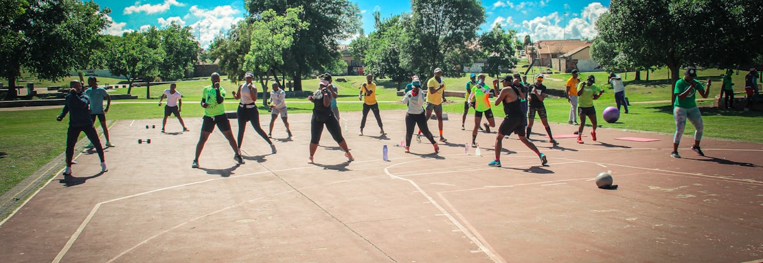 A group of people are exercising outdoors on a concrete court, surrounded by green spaces and trees. They are standing in a formation, appearing to engage in an organized fitness class. Each participant wears athletic clothing, and several carry exercise equipment such as dumbbells and ropes. The sky is bright blue with scattered fluffy clouds, creating a vibrant and energetic atmosphere.