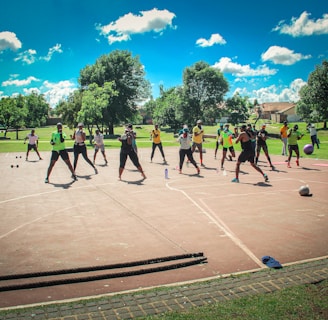 A group of people are exercising outdoors on a concrete court, surrounded by green spaces and trees. They are standing in a formation, appearing to engage in an organized fitness class. Each participant wears athletic clothing, and several carry exercise equipment such as dumbbells and ropes. The sky is bright blue with scattered fluffy clouds, creating a vibrant and energetic atmosphere.