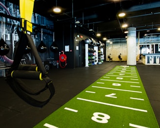 A modern gym space featuring a green track laid out on the floor with numbered lanes. TRX suspension trainers hang prominently in the foreground. The gym is equipped with various exercise equipment, including kettlebells and punching bags. In the background, there is a FIT sign near a reception desk under a low-ceilinged industrial design.