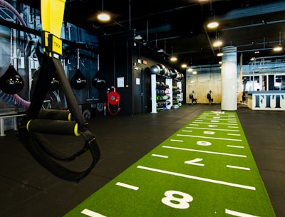A modern gym space featuring a green track laid out on the floor with numbered lanes. TRX suspension trainers hang prominently in the foreground. The gym is equipped with various exercise equipment, including kettlebells and punching bags. In the background, there is a FIT sign near a reception desk under a low-ceilinged industrial design.