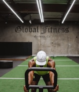 A person in athletic wear is pushing a sled on green turf inside a dimly lit gym with 'Global Fitness' written on the wall. The setting includes exercise equipment and industrial elements, such as visible metal beams and a rolled-up garage door.