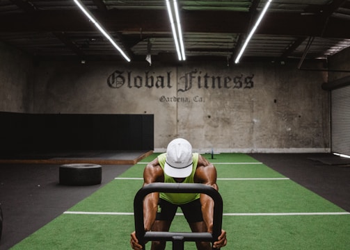 A person in athletic wear is pushing a sled on green turf inside a dimly lit gym with 'Global Fitness' written on the wall. The setting includes exercise equipment and industrial elements, such as visible metal beams and a rolled-up garage door.