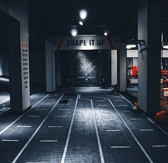 A dimly lit indoor gym with a central pathway marked with lane lines. The ceiling features multiple lights, and there are exercise equipment and motivational signage, including one that reads 'SHAPE IT UP'. The ambiance suggests a focus area for fitness activities.