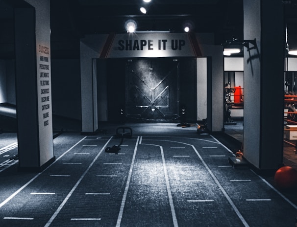 A dimly lit indoor gym with a central pathway marked with lane lines. The ceiling features multiple lights, and there are exercise equipment and motivational signage, including one that reads 'SHAPE IT UP'. The ambiance suggests a focus area for fitness activities.