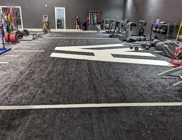 A spacious gym with a black and white color scheme featuring various fitness equipment. On the left, numerous weight plates and bars are organized on racks. In the center, a large open workout area is visible with a textured floor and white directional markings. To the right, there are treadmills, stationary bikes, and other workout machines along with large exercise balls. Exercise bands and weightlifting belts are hanging on hooks at the back wall. A single person is seen near a door, adding a human element to the scene.