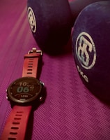 A close-up view of a fitness watch with a red strap placed on a textured purple exercise mat. Beside the watch are two large blue dumbbells with white branding on the ends.