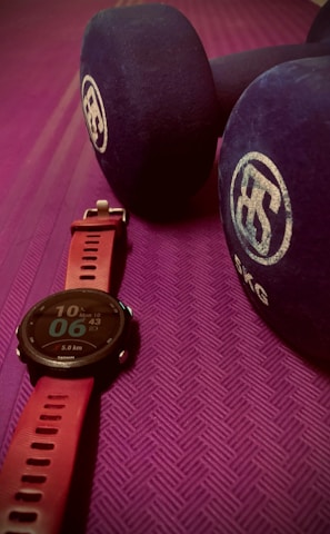 A close-up view of a fitness watch with a red strap placed on a textured purple exercise mat. Beside the watch are two large blue dumbbells with white branding on the ends.