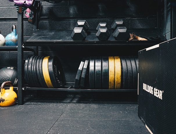 A gym setting featuring various fitness equipment, including kettlebells, stacked weight plates on a rack, dumbbells, and a medicine ball. The gym has a rugged, industrial vibe with black walls and a Bulldog Gear plyo box.