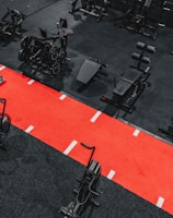 A gym setting featuring a variety of exercise equipment such as weight machines, stationary bikes, and a sled on a bright red track. The gym has a modern aesthetic with black and red color schemes.