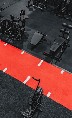 A gym setting featuring a variety of exercise equipment such as weight machines, stationary bikes, and a sled on a bright red track. The gym has a modern aesthetic with black and red color schemes.