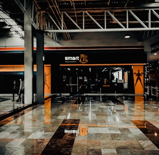 A fitness center entrance with the branding 'smart fit' illuminated above the doors. The interior has a modern design, featuring dark glass and bright orange accents. A reflective marble floor extends in front, capturing reflections from the surroundings. There are two individuals standing and walking towards the left.