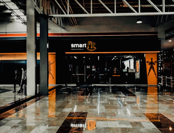 A fitness center entrance with the branding 'smart fit' illuminated above the doors. The interior has a modern design, featuring dark glass and bright orange accents. A reflective marble floor extends in front, capturing reflections from the surroundings. There are two individuals standing and walking towards the left.